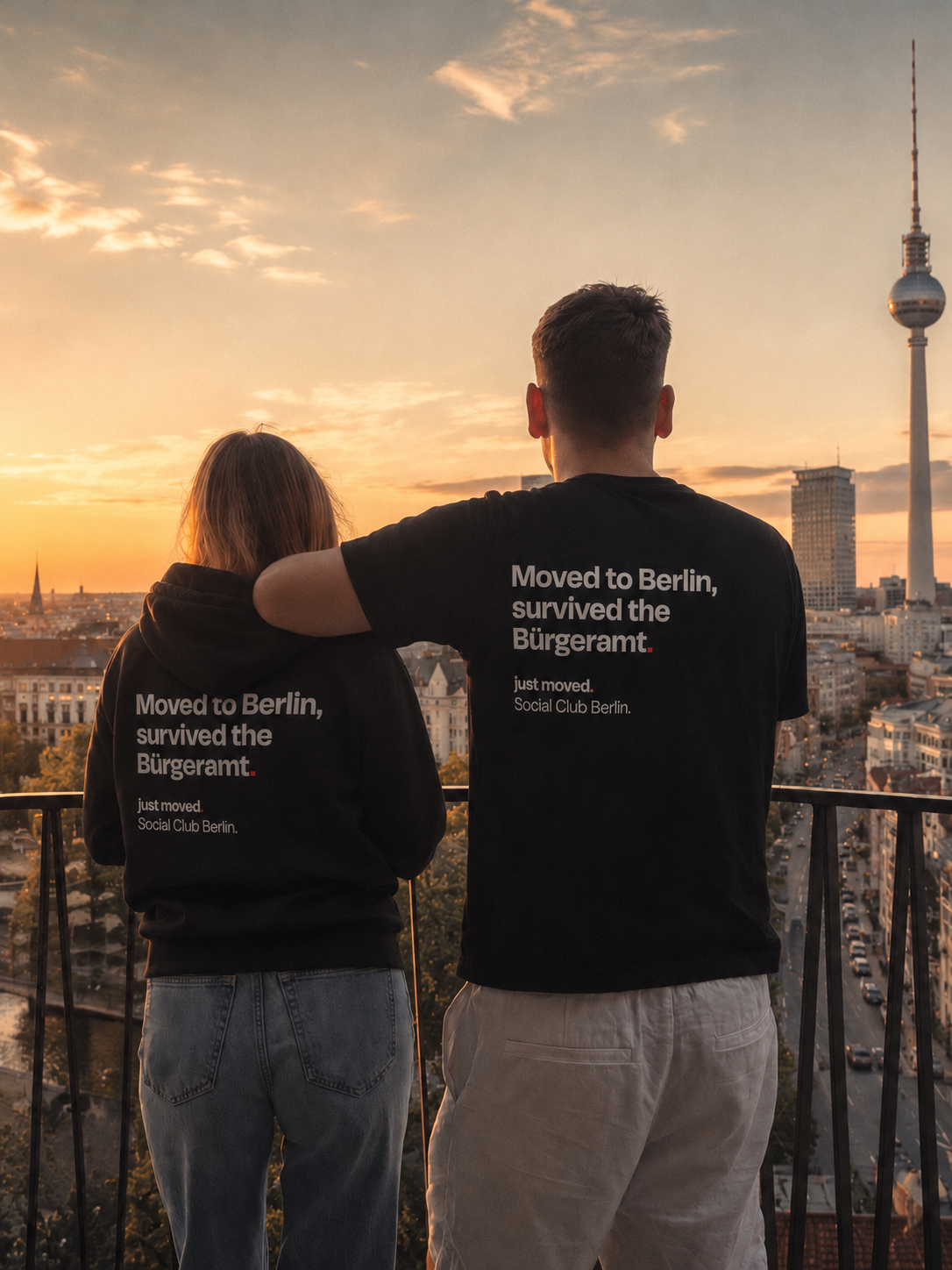 Alex and Vanessa on a Berlin rooftop at sunset, wearing the justmoved club tees that read 'Moved to Berlin, survived the Bürgeramt.'